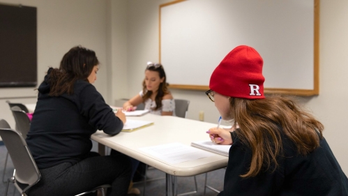 Three students study around a table. One student is wearing a red Rutgers hat