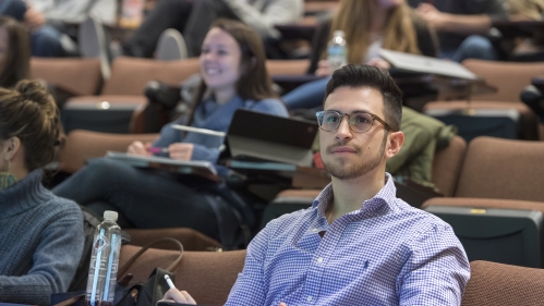 A student listens to a lecture in a lecture hall, other students are blurred in the background