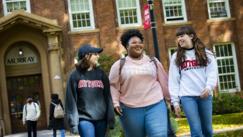 Three students wearing Rutgers sweatshirts and backpacks walk outside Murray Hall on the College Avenue campus