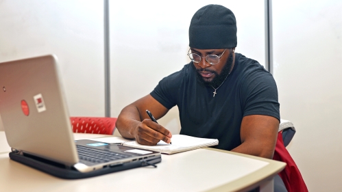 A student wearing a black t-shirt and hat writes in a notebook in the learning centers.