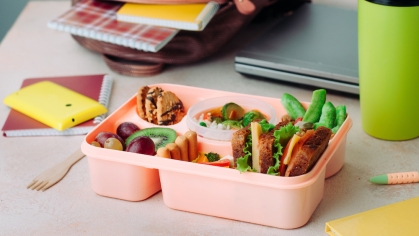 A pink bento box with food sits on a desk beside a green water bottle, a laptop and school supplies.