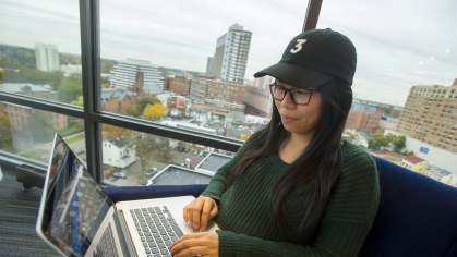 A student wearing a green sweater and a black baseball hat works on her laptop in the 13th floor Sojourner Truth apartments Sky Lounge overlooking the city of New Brunswick. 