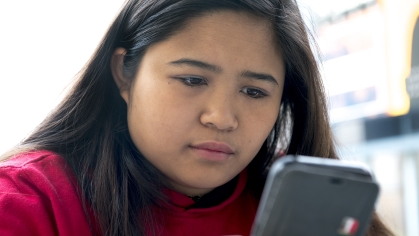 A student with long brown hair and a red sweatshirt reads her cell phone