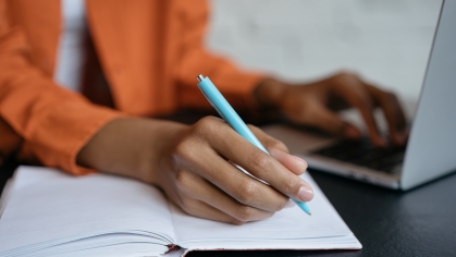A student wearing an orange shirt writes in a notebook and works on a laptop