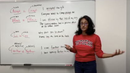 A writing coach in a red sweater stands in front of a white board while teaching