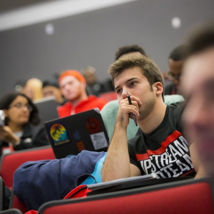 A student listens to a lecture in the academic building lecture hall, other students are blurred in the foreground and background