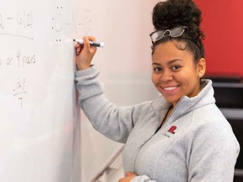 A student wearing a gray Rutgers sweater writes on a dry-erase board in the Learning Centers