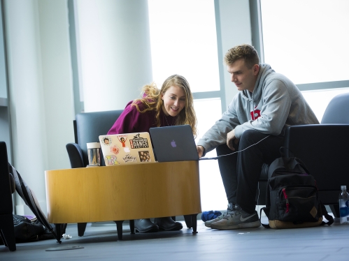 Two students study on their laptops in a hallway lounge