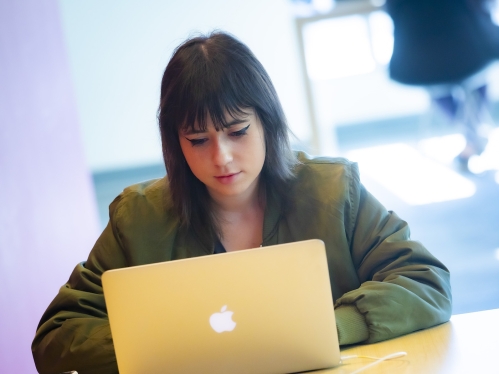A student with brown hair wearing a green jacket studies on her laptop in the academic building