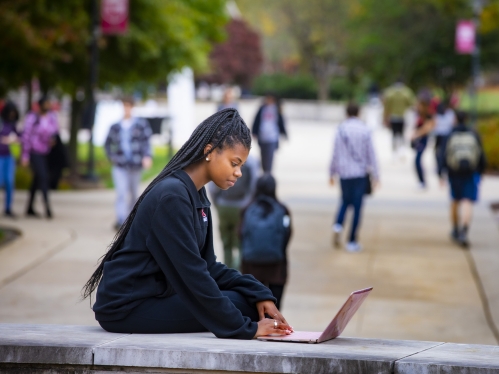 A student with long braids and a black sweatshirt studies on her laptop while sitting on a brick wall on Livingston campus